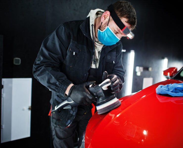 Car detailing concept. Man in face mask with orbital polisher in repair shop polishing orange suv car.