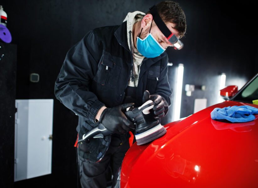 Car detailing concept. Man in face mask with orbital polisher in repair shop polishing orange suv car.